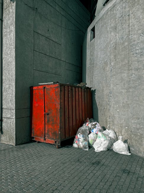 A middle-aged man with short dark hair and light skin is seen bending over to dispose of rubbish into a silver, cylindrical public litter bin with an open-top design. He is wearing a black T-shirt wit