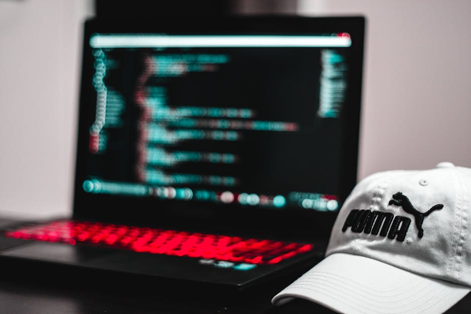 A close-up view of a person's hands typing on a laptop keyboard placed on a dark surface. The laptop screen displays lines of code in multiple colors, including green, blue, and white, on a dark backg
