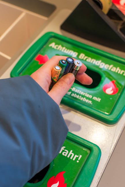 A person's hand, wearing a blue sleeve, is seen inserting used batteries into a green recycling container designated for battery disposal. The container has a rectangular opening and features German t