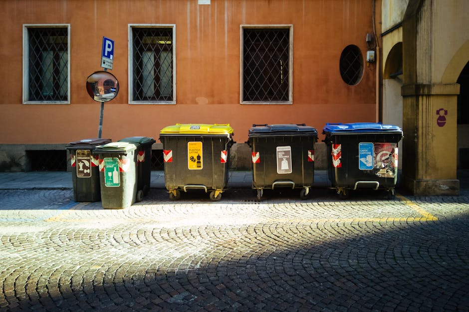 A person's hand, wearing a blue sleeve, is seen inserting used batteries into a green recycling container designated for battery disposal. The container has a rectangular opening and features German t