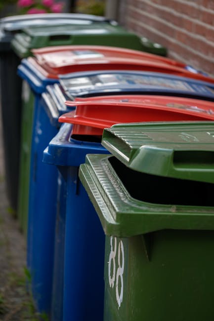 A row of multiple large wheelie bins lined up outdoors along a paved pathway adjacent to a brick wall. The bins are made of durable plastic with ribbed lids, featuring different colours including gree